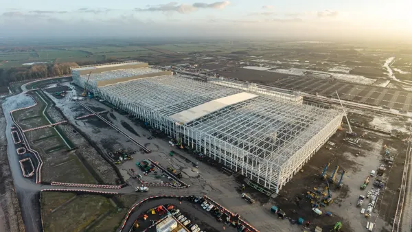A skyshot of a large steel-framed structure that sits in a large field during twilight. Construction equipment dots the landscape around it.