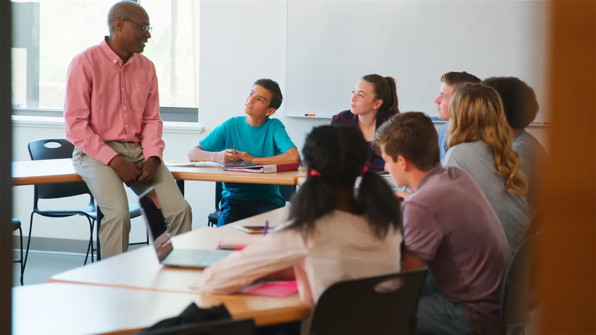An adult is leaning against a desk in a classroom. A handful of students are seated at desks looking at the adult.