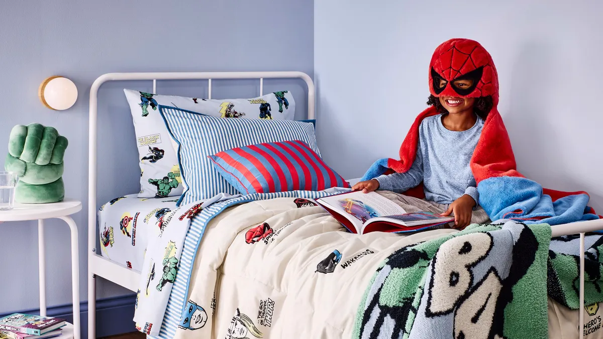 A child sitting on a Marvel-themed bed wearing a blanket with a Spiderman mask.