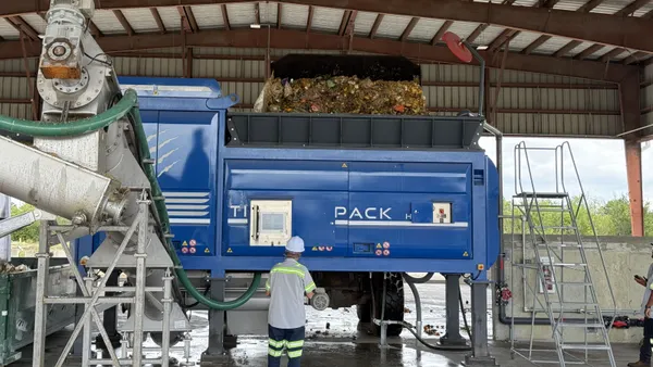 A man stands in an industrial shed in front of a large machine with a hopper feeding in food waste for processing.