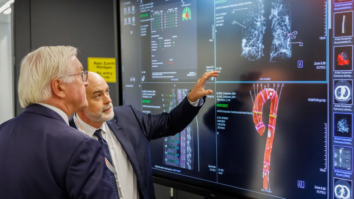 Two people dressed in business attire look at a large monitor with four dived screens displayed, and one person points with their hand.