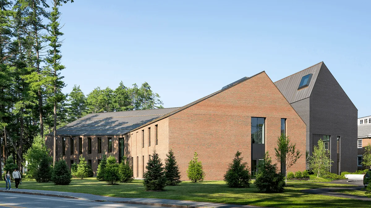 Two buildings with non-standard roof shapes rise above a grassy, rolling hill during a day with blue sky. The buildings are surrounded by green space and trees, and look very modern.