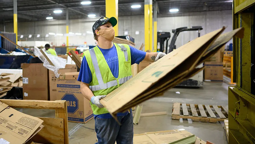 An employee, wearing a beig face mask and a yellow vest uses a cardboard baler.