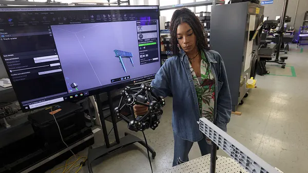 A woman holds a 3-D lab apparatus in her right hand. A screen is behind her depicting as aspect of a 3-D model.