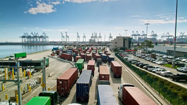 Drone shot of the front gate of a container terminal at the Port of Los Angeles on a sunny day. Trucks are lined up to leave and enter, and in the background massive container ships are sitting in their berths being loaded and unloaded by cranes.