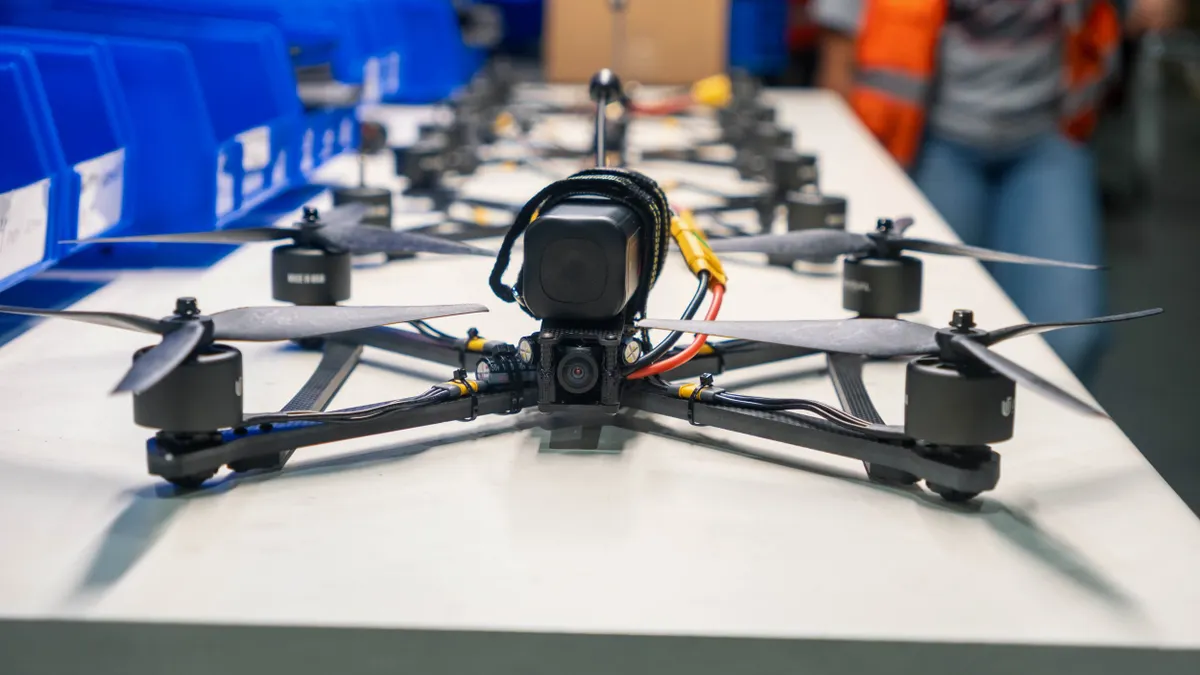 A row of black drones lined up on a table next to blue plastic shelf bins.