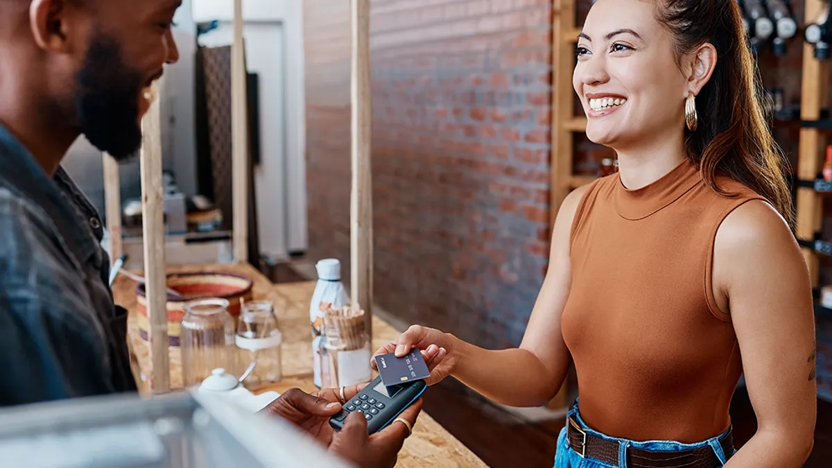 A woman smiles while holding her phone over a payment terminal held by a man in a cafe. The setting is bright and welcoming with a brick wall backdrop.