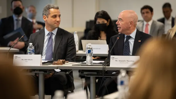 Rows of people sit at tables with name cards in front of each of them. The two people in the foreground look at each other as they converse.