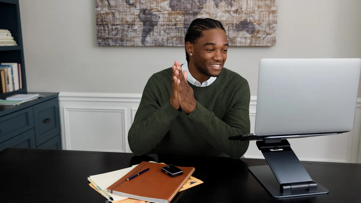 A person sits at a desk, smiling, with a square device on top of a stack of notebooks. They are looking at a laptop.