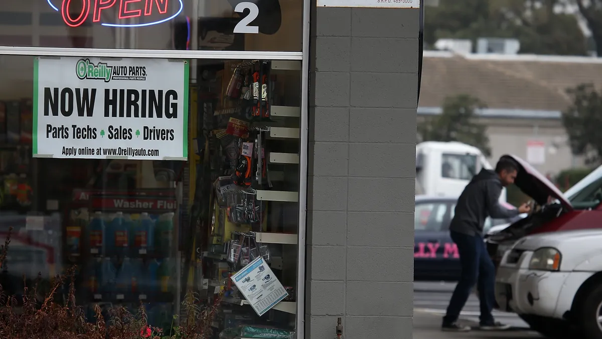 image of an open O'Reilly's store front with a help wanted sign in the window and a man working on his car to the right.