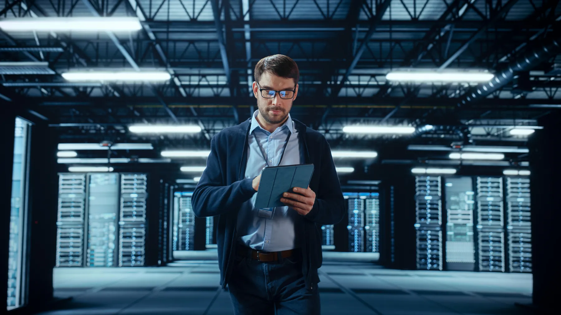 Male IT Specialist Walks Between Row of Operational Server Racks in Data Center. Engineer Uses Tablet Computer for Maintenance.