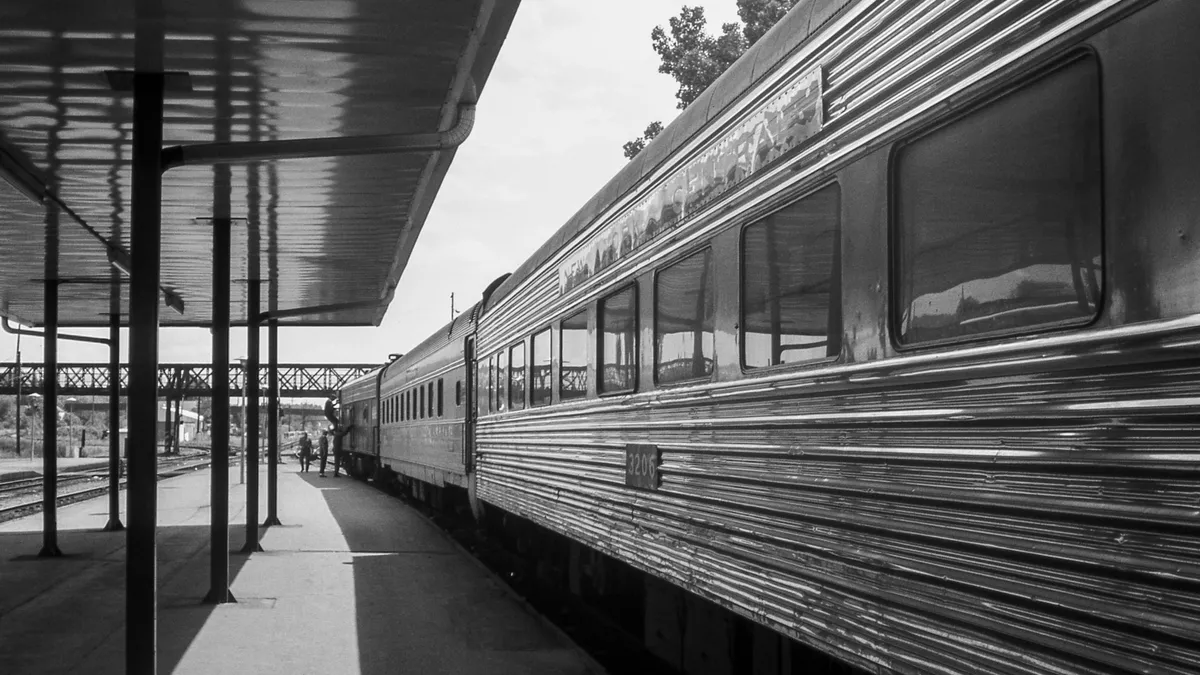 A black and white photo of an old streamliner passenger train stopped at a station with a view toward the front of the train and several railroad workers alongside.