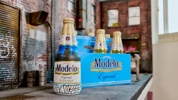 A painted bottle of Modelo sits on a table infront of a blue six pack which is missing a bear. The background is blurred and a paint palette sits in the foreground.
