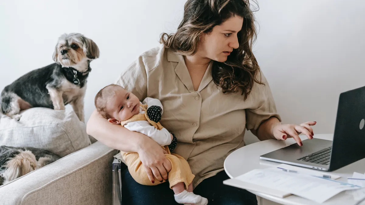 A parent types at a laptop, holding a baby