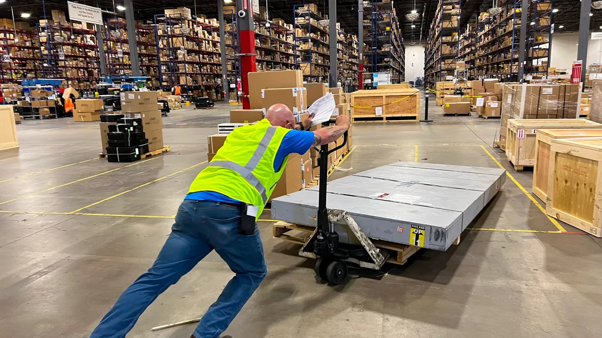 A person wearing a neon green worker vest pushes a large flat shipment on a pallet jack into a warehouse with rows of supply.