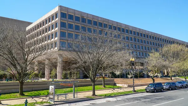A large modern government office building with a grid of square windows, in front, leafless trees line the sidewalk, and a bus stop with a shelter. 