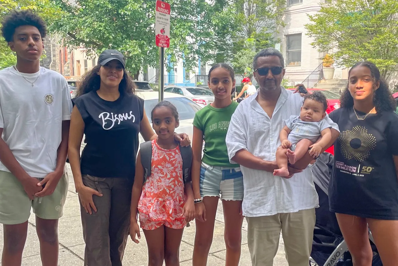 A family of adults and children stand on a sidewalk facing the camera.