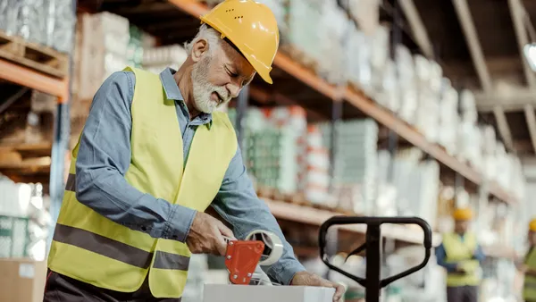 Senior male worker packing cardboard box with tape gun dispenser in warehouse