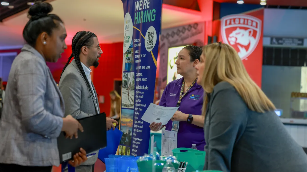 Individuals speak across a table with a "we're hiring" sign in the background.