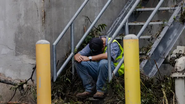 A person wearing a vest and boots sits with head bowed on steps amid debris.