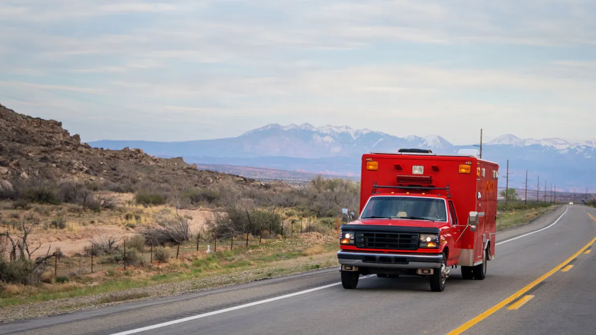 A red ambulance drives on a road with mountains in the background.