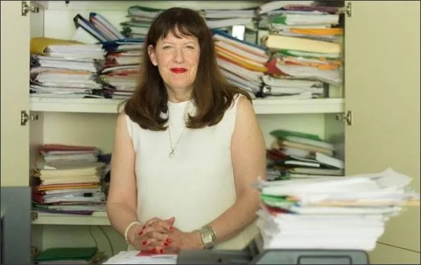 A profile photo of an adult looking at the camera. Behind the adult is a bookshelf of papers and books.