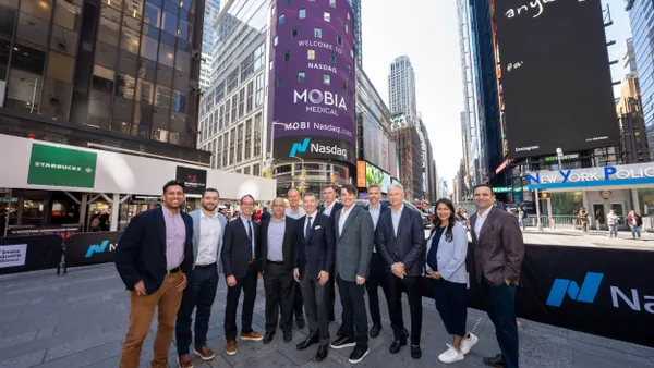 A group of people stands in Times Square in front of a large ad thaqt says "Welcome to NASDAQ Mobia Medical."