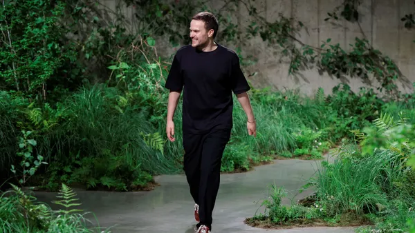 A person in a black top and pants walks down a runway that features greenery along the sides.