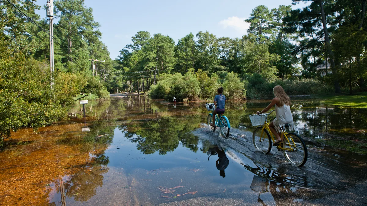 two girls ride bikes away from the viewer on a tree-lined street partially covered with water from flooding.