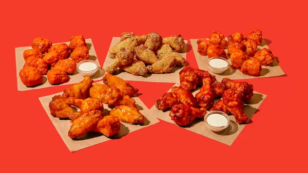 A photo of five different flavors of wings sitting on squares of brown paper against a red background. There are small cups of ranch dipping sauce near three of the types of wings.