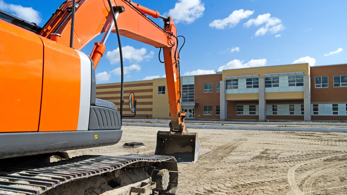 An orange excavator is in the forefront. In the background is a school under construction