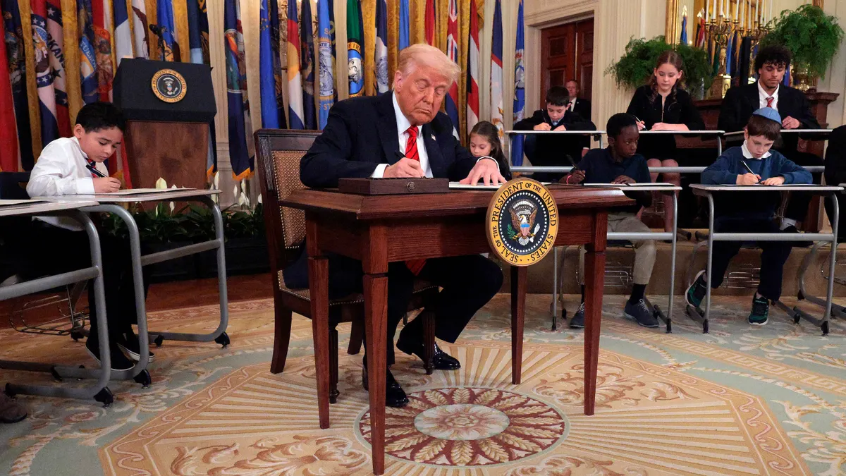 An adult sits at a small desk on a stage in a room, holding a pen and writing on a paper. Rows of children are sitting at individual desks behind the adult.