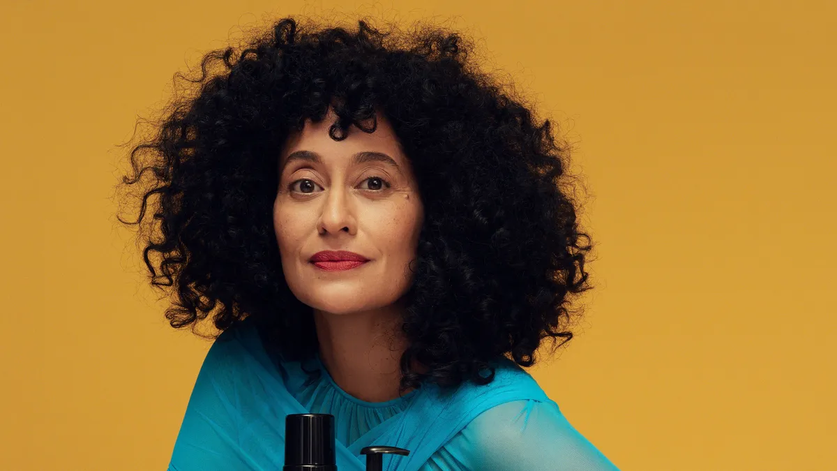 Tracee Ellis Ross sits in a blue dress behind a lineup of her haircare.