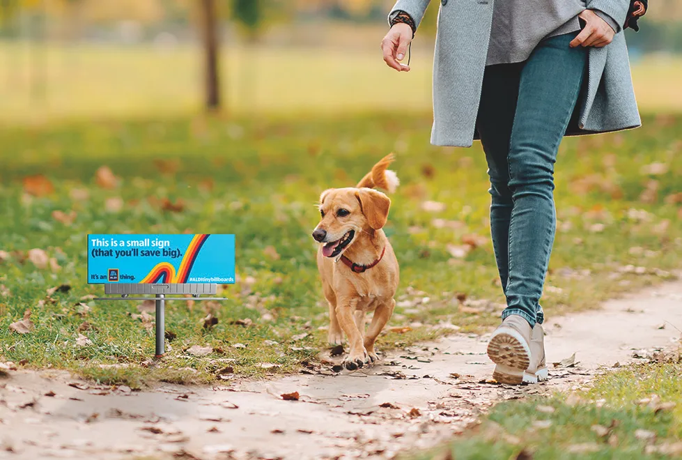 A person and dog walk by a miniature yard sign that says "This is a small sign that you'll save big. It's an Aldi thing."