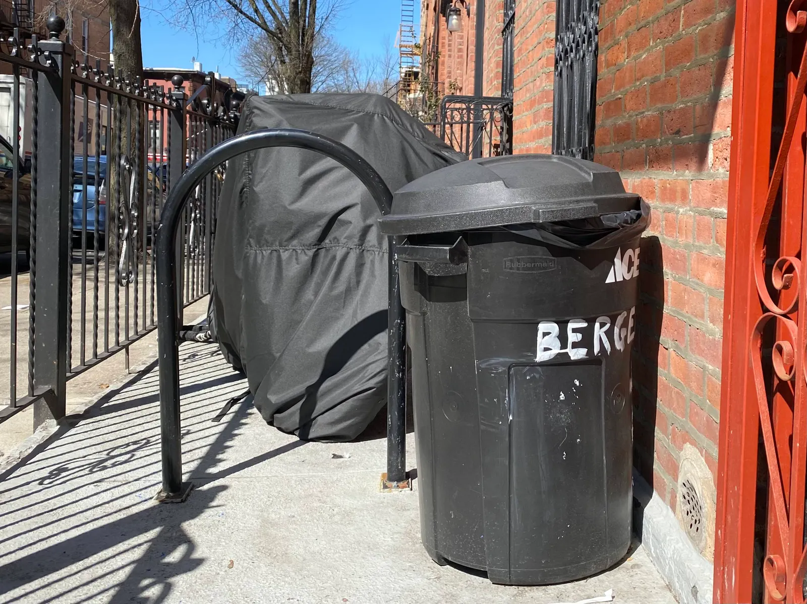 A black lidded trash can sits in front of a building in a fenced-off area. Next to it are a bike rack and covered moped.