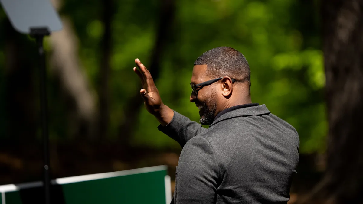 A man wearing a dark suit and sunglasses stands and waves in a forested area.