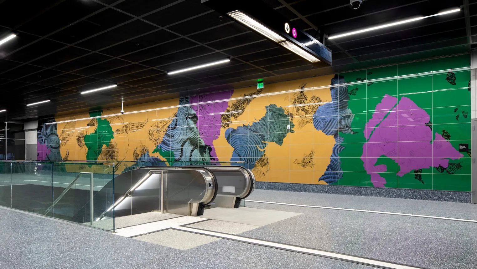 Wall art in colors of yellow, green purple and blue behind an underground train platform and escalator.