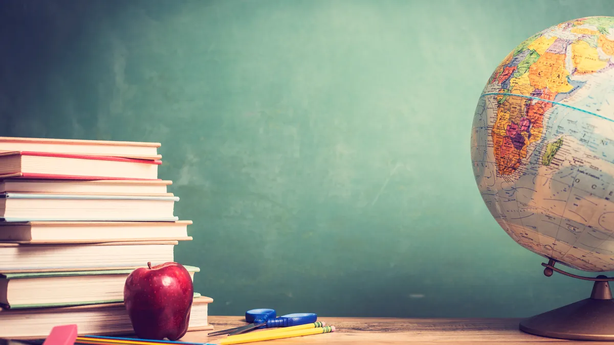 A stack of books on a table sits next to a red apple and globe. In the background is a chalk board