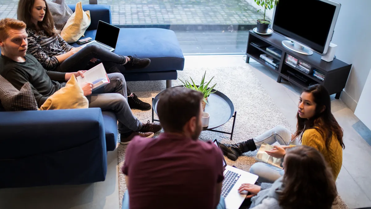 People sit on couches in a living room, holding books and laptops.