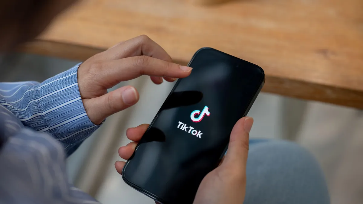 A person holding a smartphone with the TikTok splash screen visible, seated at a table with a hand about to tap the screen.