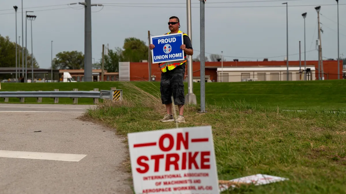 A blurred red and white "On strike" sign on top of grass, and a man in the background holding a white and blue "Proud union home" sign.
