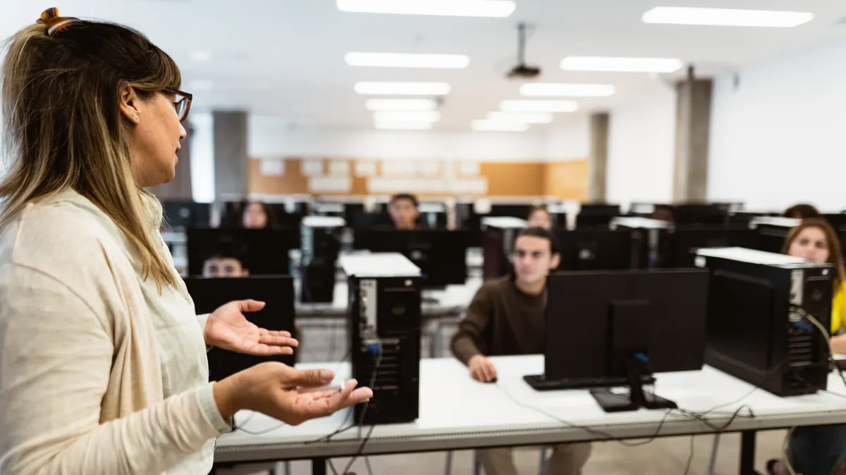 A teacher lectures students at desks with computers.