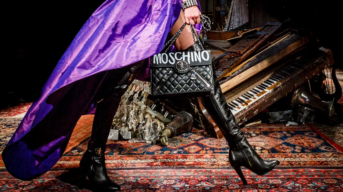 A model walks the runway at the Moschino fashion show wearing a purple high-low skirt and knee-high black boots and carrying a Moschino bag.