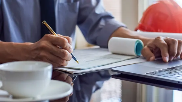A man sits at a desk with a notepad and a hard hat.