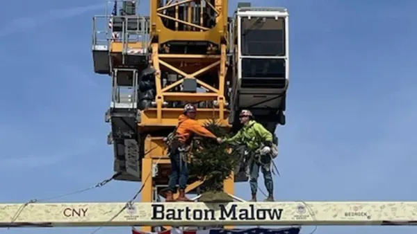 Workers in front of heavy equipment stand with a beam that reads "Barton Malow" on top of a tall building.