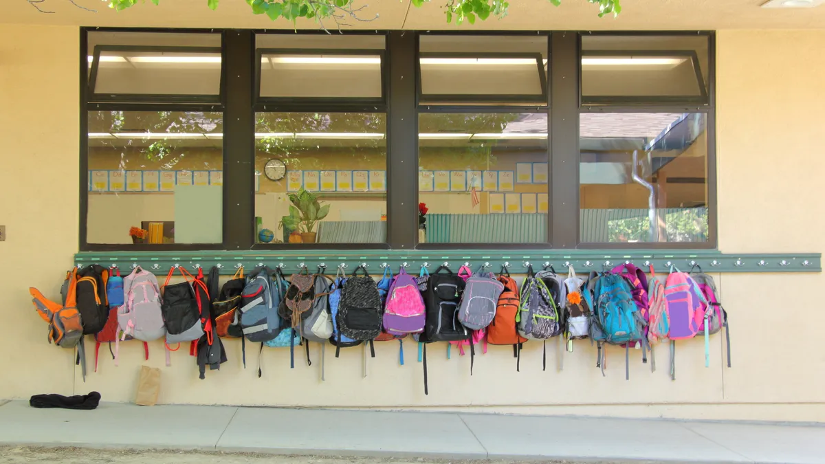 Students' backpacks hang in a row under a window outside a school room.