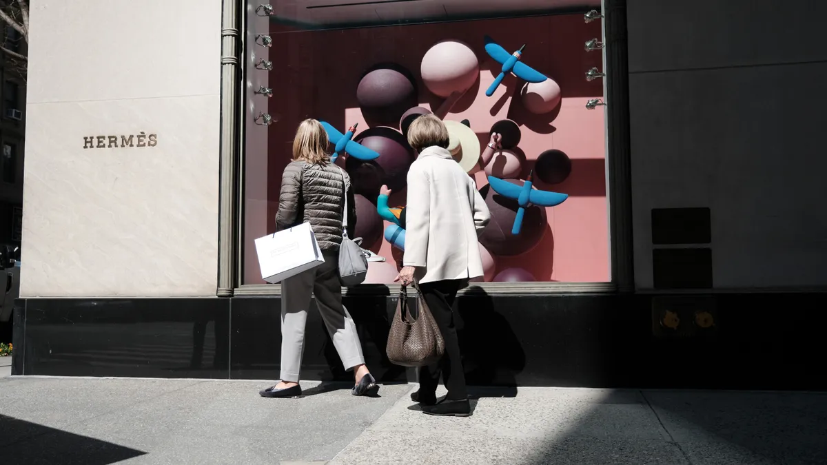 People walk by luxury retail store Hermes on a sunny day, holding shopping bags and looking in the window.