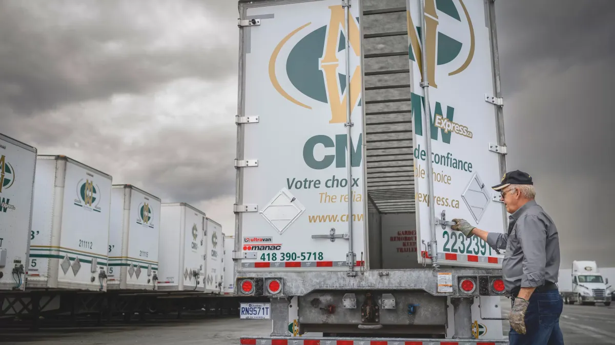 A driver stands near an open CMW Express trailer surrounded by other trailers in a company parking lot.