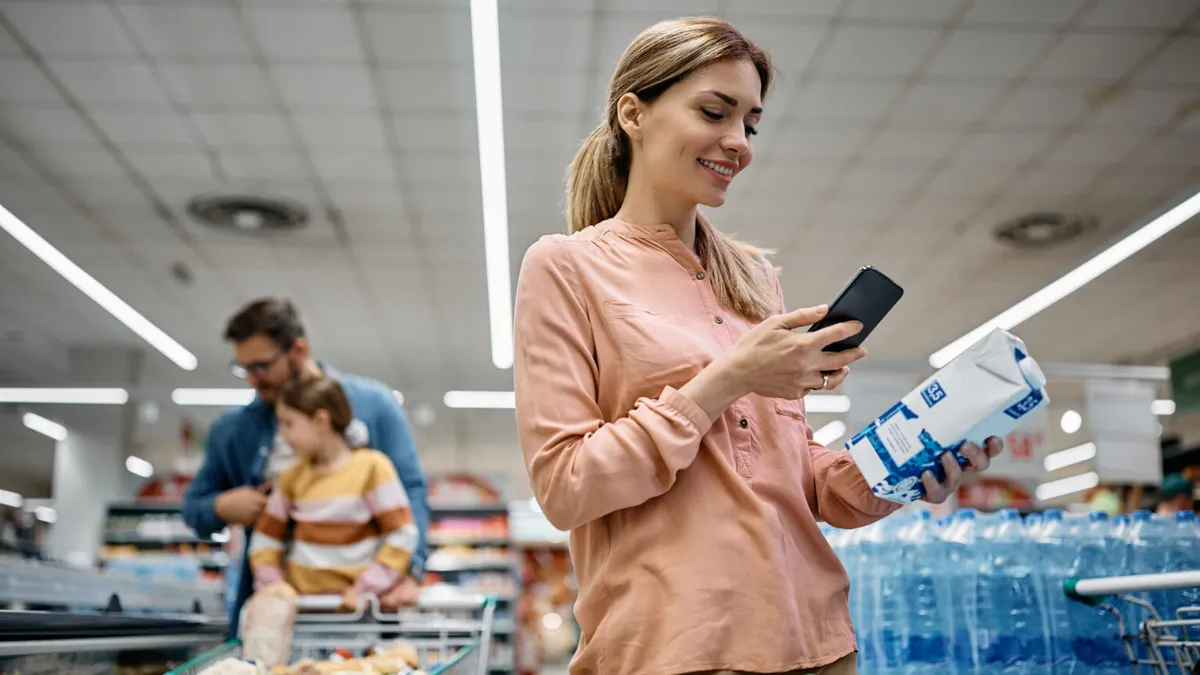 A woman in a supermarket smiles and holds a smartphone above a carton