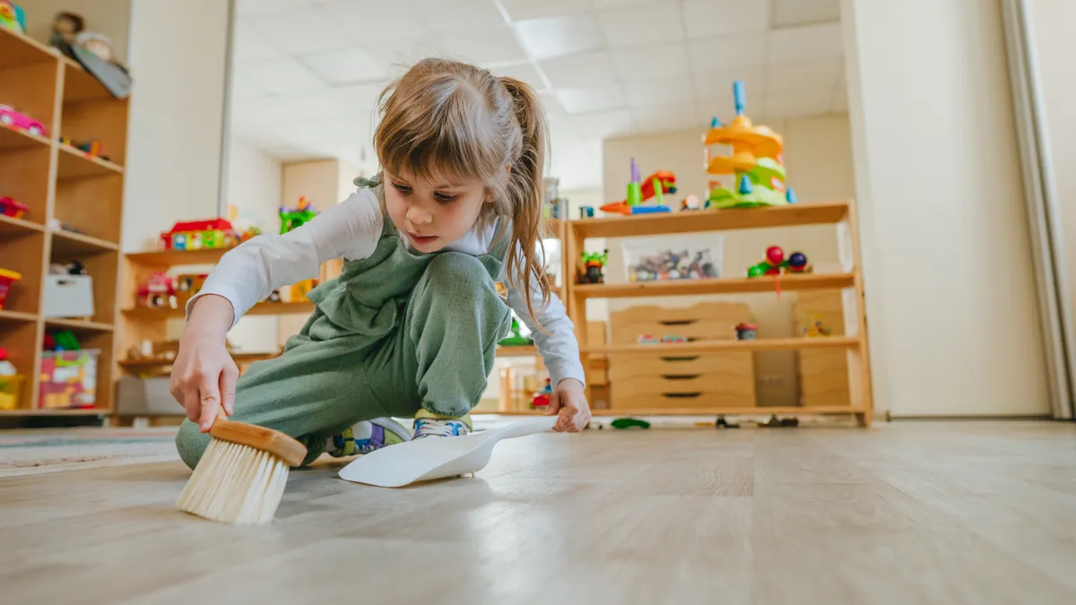 A young child kneels on the floor in a classroom and uses a handheld broom and dustpan to sweep the floor.